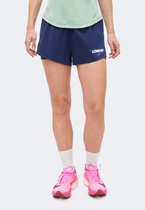 Person iført marineblå shorts med teksten "LONDON", lys farvet top, hvide sokker og lyserøde løbesko, der står på en hvid baggrund.