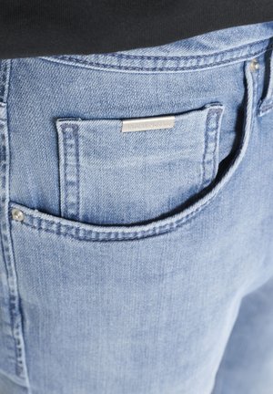 Tasca di jeans in denim azzurro chiaro con etichetta metallica del marchio sulla piccola tasca interna e cuciture visibili lungo i bordi.