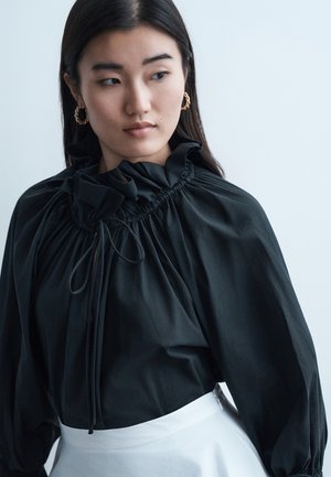 Γυναίκα με μακριά σκούρα μαλλιά, που φοράει χρυσούς κρίκους στα αυτιά, μια μαύρη μπλούζα με βολάν και κορδόνια, και μια λευκή φούστα, κοιτάζοντας προς τα αριστερά της.