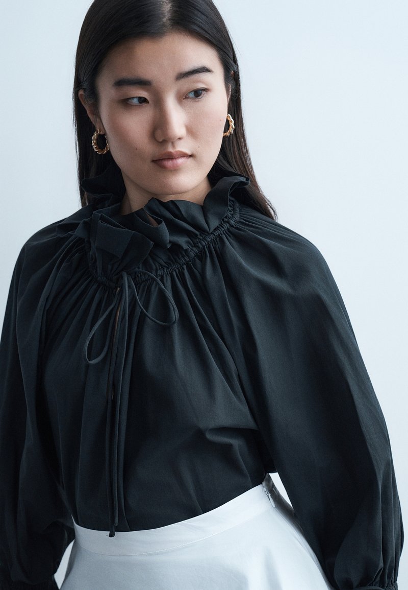 Γυναίκα με μακριά σκούρα μαλλιά, που φοράει χρυσούς κρίκους στα αυτιά, μια μαύρη μπλούζα με βολάν και κορδόνια, και μια λευκή φούστα, κοιτάζοντας προς τα αριστερά της.