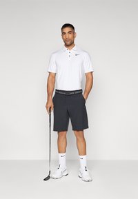 Nike golf outfit bestående af en hvid kortærmet polo, sorte shorts, sort bælte med grå detaljer og hvide sko med matchende sokker, mens han holder en golfkølle.
