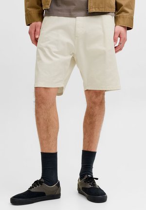 Homme portant un short beige, des chaussettes noires et des baskets noires, debout les mains détendues le long du corps sur un fond blanc.