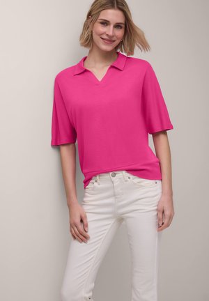 Smilende kvinde med kort blondt hår iført en lyserød kortærmet top og hvide tætsiddende jeans mod en lys baggrund.