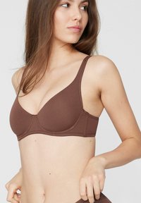Hnědá bezešvá podprsenka s nastavitelnými ramínky, tvarovanými košíčky a měkkou pružnou látkou. Vyznačuje se hladkou texturou a jednoduchým designem.