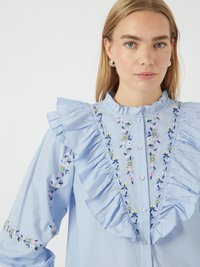 Camicia button-up azzurro chiaro con colletto e maniche arricciate, decorata con ricami floreali rosa, blu e verdi sul davanti e sulle maniche.