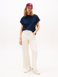 Top blu navy a maniche corte con dettagli a bottone, pantaloni beige a gamba larga e sneakers bianche, abbinati a un cappellino borgogna con logo.