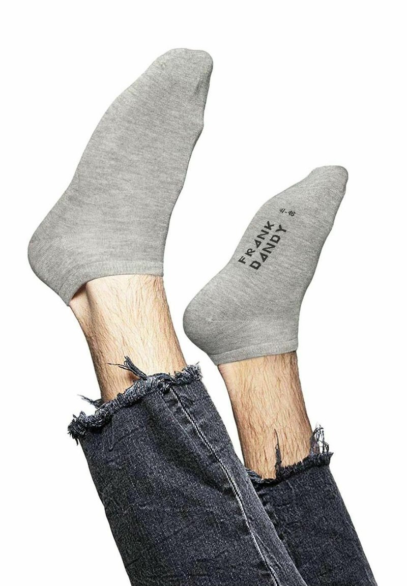 Calcetines tobilleros de algodón gris con textura suave, exhibidos en pies que sobresalen de pantalones vaqueros oscuros desgastados, con el nombre de la marca impreso en un lateral.