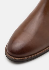 Hnědá kožená kotníková bota s hladkou texturou, špičatou špičkou a jemným prošíváním podél podešve. Klasický design a bohatá barva.