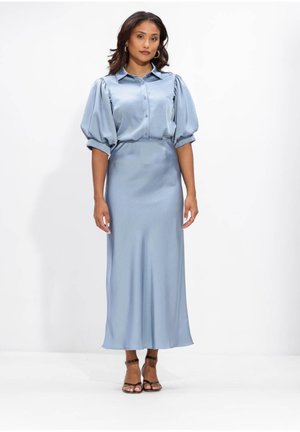 Vrouw die een lichtblauwe satijnen blouse met pofmouwen draagt en een bijpassende hooggesloten rok tot op de enkels, staand tegen een effen witte achtergrond.