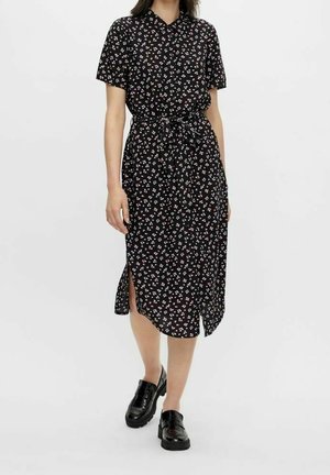 Frau in einem schwarzen Midi-Kleid mit kleinen weißen und rosa Blumenmuster, kurzen Ärmeln, Kragen, seitlichem Schlitz und schwarzen klobigen Schuhen.