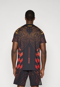 Μαύρο αθλητικό πουκάμισο με κόκκινο και πορτοκαλί σχέδιο φτερωτών γραφικών, με κοντά μανίκια και λογότυπο Venum στο γιακά. Συνδυάζεται με αντίστοιχα σορτς.