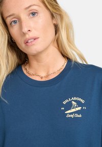 Mørkeblå T-shirt med rund hals, udsmykket med et elfenbensfarvet logo og design på venstre bryst, der viser en surfer og teksten "Surf Club."