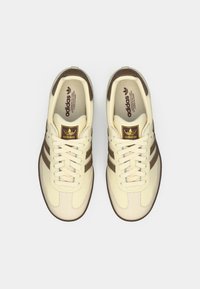 Buty sportowe Adidas z kremową skórzaną cholewką, brązowymi zamszowymi paskami i złotym logo. Okrągły nosek z teksturowaną kremową gumową podeszwą.