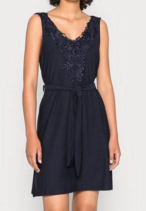 Robe bleu marine sans manches avec un col brodé de motifs floraux et une ceinture en tissu nouée à la taille, portée par une personne debout devant un fond uni.