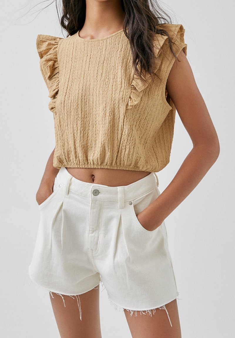 Femme portant un haut beige texturé sans manches avec des épaules à volants et un short en denim blanc taille haute avec des ourlets effilochés.