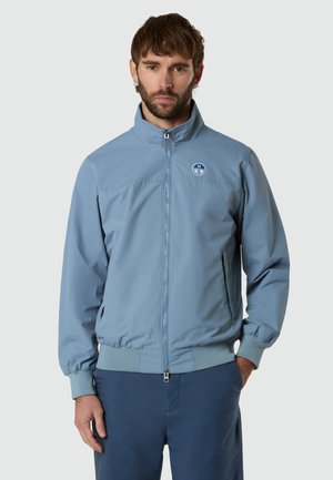 Mann mit Bart, der eine hellblaue Zip-up-Jacke mit Kompasslogo auf der Brust trägt und vor einem schlichten hellen Hintergrund steht.