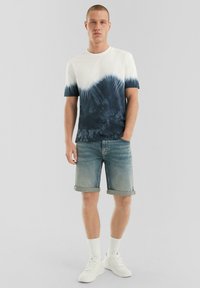 Weißes und marineblaues Batik-T-Shirt mit kurzen Ärmeln, kombiniert mit hochgekrempelten Jeansshorts. Modell trägt weiße Sneakers und knöchellange weiße Socken.