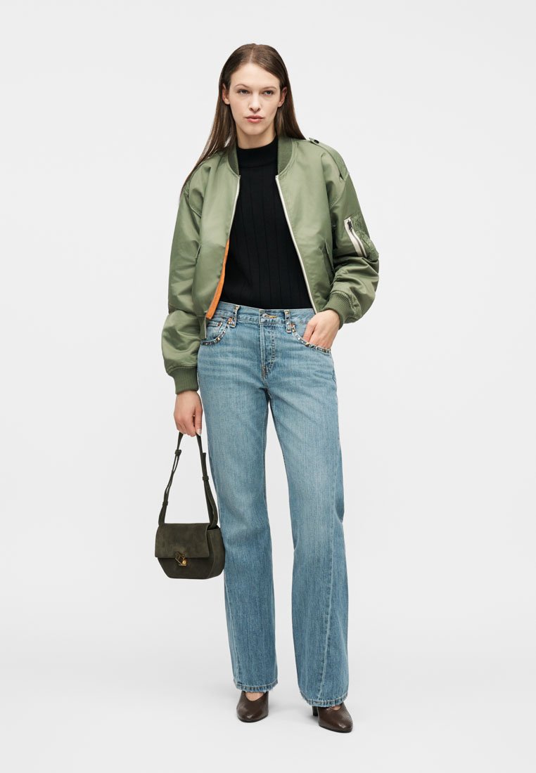 Femme portant une veste bomber verte, un haut noir, un jean bleu clair, des chaussures marron, tenant un petit sac à main foncé, debout devant un fond blanc.