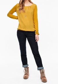 Senapsgul långärmad topp med rund halsringning och veck, i kombination med mörkblå denimjeans, uppvikta ärmslut och bruna snörstövlar.