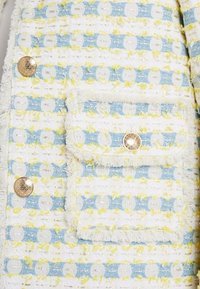 Gewebter Stoff in Blau, Weiß und Gelb, mit einem strukturierten Muster, goldenen Knöpfen und einer Klapptasche mit fransigen Kanten.