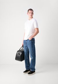 Witte katoenen T-shirt, relaxte blauwe spijkerbroek, zwarte leren tas en zwarte sneakers met witte zolen, alles tegen een effen achtergrond.
