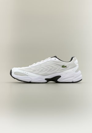 Biały sportowy sneaker z czarnymi akcentami na podeszwie, teksturowaną cholewką, sznurowanym designem i zielonym logo krokodyla na szarym tle.