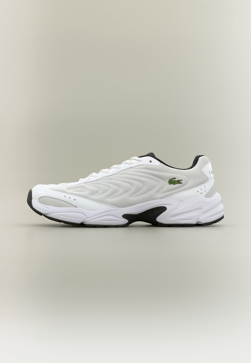 Baskets de sport blanches avec des accents noirs sur la semelle, dessus texturé, design à lacets et logo crocodile vert sur fond gris.