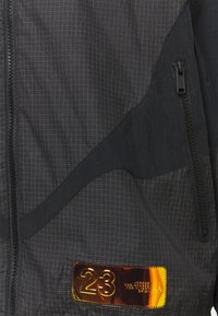 Zwarte jas met ruitpatroon, met een glanzend, metalen label waarop "23" is gegraveerd, en contrasterende, gestructureerde panelen en zakken met rits.