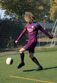 Bordo zestaw piłkarski z turkusowymi akcentami, długimi rękawami i okrągłym dekoltem. Gracz w ruchu kopie czarno-białą piłkę na zielonej murawie.