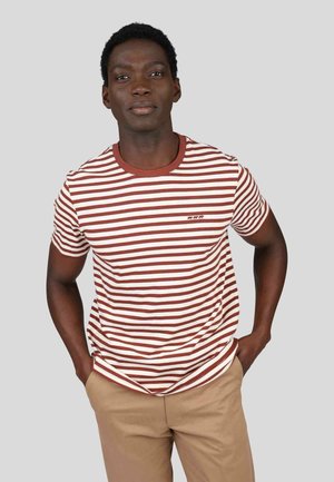 T-shirt rayé en blanc et bordeaux, à encolure ronde, à manches courtes, avec un petit logo brodé sur la poitrine. Porté avec un pantalon kaki.