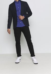 Conjunto masculino que incluye un blazer marino, una camisa azul con lunares, pantalones negros y zapatillas Nike blancas con detalles en negro. Diseño simple y moderno.