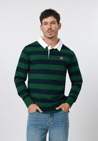 Άνδρας που φοράει μακρυμάνικη πόλο μπλούζα με πράσινες και ναυτικές ρίγες, λευκό γιακά και ανοιχτό μπλε τζιν, με τα χέρια στις τσέπες, ουδέτερο φόντο.