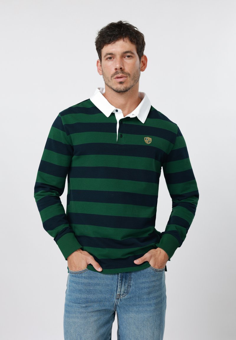 Άνδρας που φοράει μακρυμάνικ η πόλο μπλούζα με πράσινες και ναυτικές ρίγες, λευκό γιακά και ανοιχτό μπλε τζιν, με τα χέρια στις τσέπες, ουδέτερο φόντο.