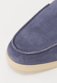 Kék velúr slip-on cipő, lekerekített orral. Jellemzője a világos bézs gumi talp és a varrás a felső élen.