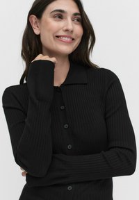 Pull en noir avec des côtes, doté d'un col et d'une fermeture à boutons sur le devant, avec des manches longues et des poignets ajustés.