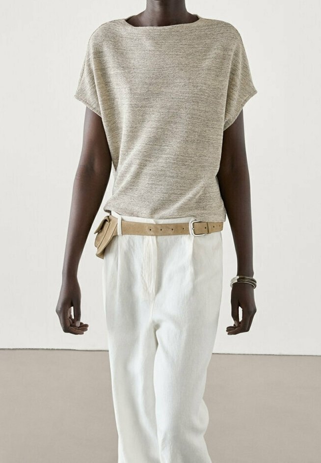 Personne à la peau foncée portant un haut beige à manches courtes en maille, un pantalon blanc taille haute, une ceinture beige avec pochette et des bracelets au poignet gauche.
