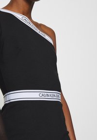 Czarna sukienka na jedno ramię z białą taśmą Calvin Klein Jeans wzdłuż klatki piersiowej i talii. Gładki, dopasowany materiał o minimalistycznym wzornictwie.