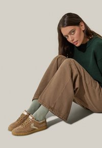 Sneaker in suede marrone con suola in gomma color gommato, caratterizzati da un accento metallico e un logo laterale. Abbinati a calzini verde chiaro e pantaloni larghi marroni.