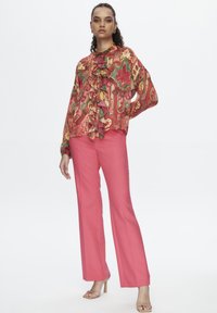 Langärmlige, rüschendige Bluse im rot-gelben Blumenmuster, kombiniert mit fuchsia-farbenen, weit geschnittenen Hosen und beigefarbenen, hohen Sandalen.