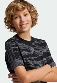 Camiseta de manga corta con estampado de camuflaje en negro y gris, con cuello redondo y tres rayas de acento en las mangas. Textura de tela suave.