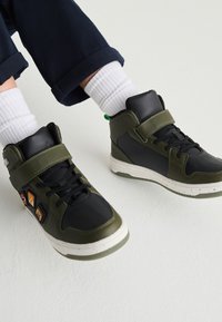Sneakers alte con schema di colori nero e verde oliva. Presentano una chiusura a cinturino, suola in gomma e toppe decorative sul lato.