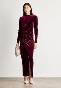 Μπορντό βελούδινο φόρεμα με μακριά μανίκια, με πτυχώσεις στη μέση και πλάγια σχισμή, συνδυασμένο με κρεμ τσάντα και μπεζ τακούνια.