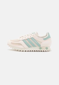 Light beige athletic sneakers with turquoise stripes, textured mesh upper, rubber sole with spike traction, and branding on the side.