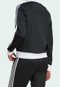 Svart sportjacka med vit krage och muddar, med tre vita vertikala ränder på ärmarna. Tillverkad av slät, elastisk tyg.