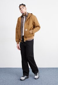 Giacca cappuccio marrone con una tasca frontale, abbinata a una camicia grigia con bottoni, pantaloni neri e scarpe da ginnastica grigie con dettagli neri.