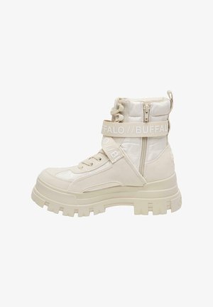 Jambes portant des bottes de combat à plateforme beige Buffalo posées sur un trottoir urbain ensoleillé près d’une rue.