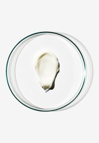 Produit blanc et crémeux en forme de goutte d'eau, sur un plat en verre transparent posé sur un fond de papier blanc. Texture lisse avec un léger éclat.