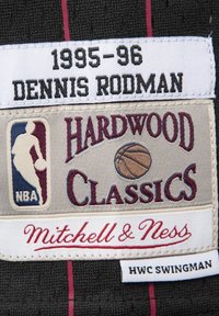 Etichetta in tessuto nera con ricami bianchi e bordeaux. Include "1995-96", "DENNIS RODMAN", "HARDWOOD CLASSICS" e "Mitchell & Ness".