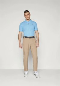 Lyseblå polo med krage, kombinert med beige bukser. Brystet med et svart belte og hvite sneakers, stående mot en nøytral bakgrunn.