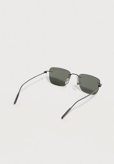 Ορθογώνια γυαλιά ηλίου χωρίς πλαίσιο με λεπτούς μεταλλικούς μαύρους βραχίονες και σκούρους φακούς σε ανοιχτό γκρι επιφάνεια.